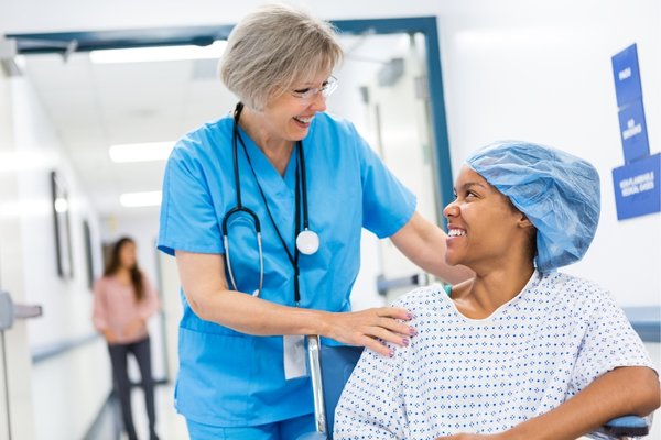 A smiling nurse in blue scrubs and a stethoscope gently places a hand on the shoulder of a patient in a hospital gown and surgical cap, seated in a wheelchair. They are in a bright, clean hospital corridor, sharing a moment of reassurance and trust.
